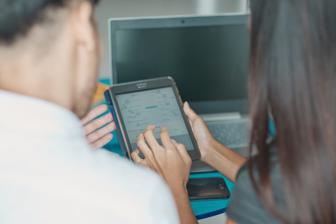 Two people look at a tablet. The view is from behind their shoulders.