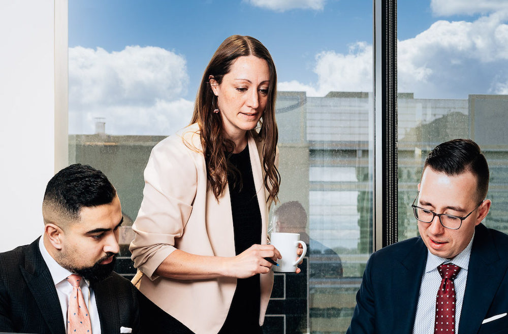 Three lawyers sit and stand around a board table